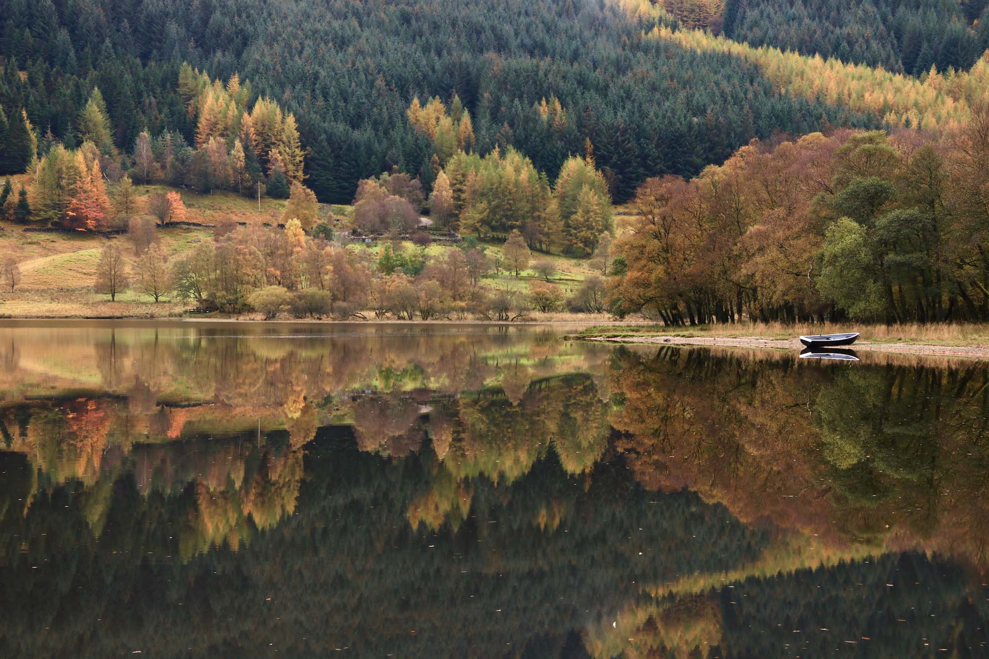 Scotland in Autumn, there is a boat in a lake which reflects the red and yellow leaves of the trees.